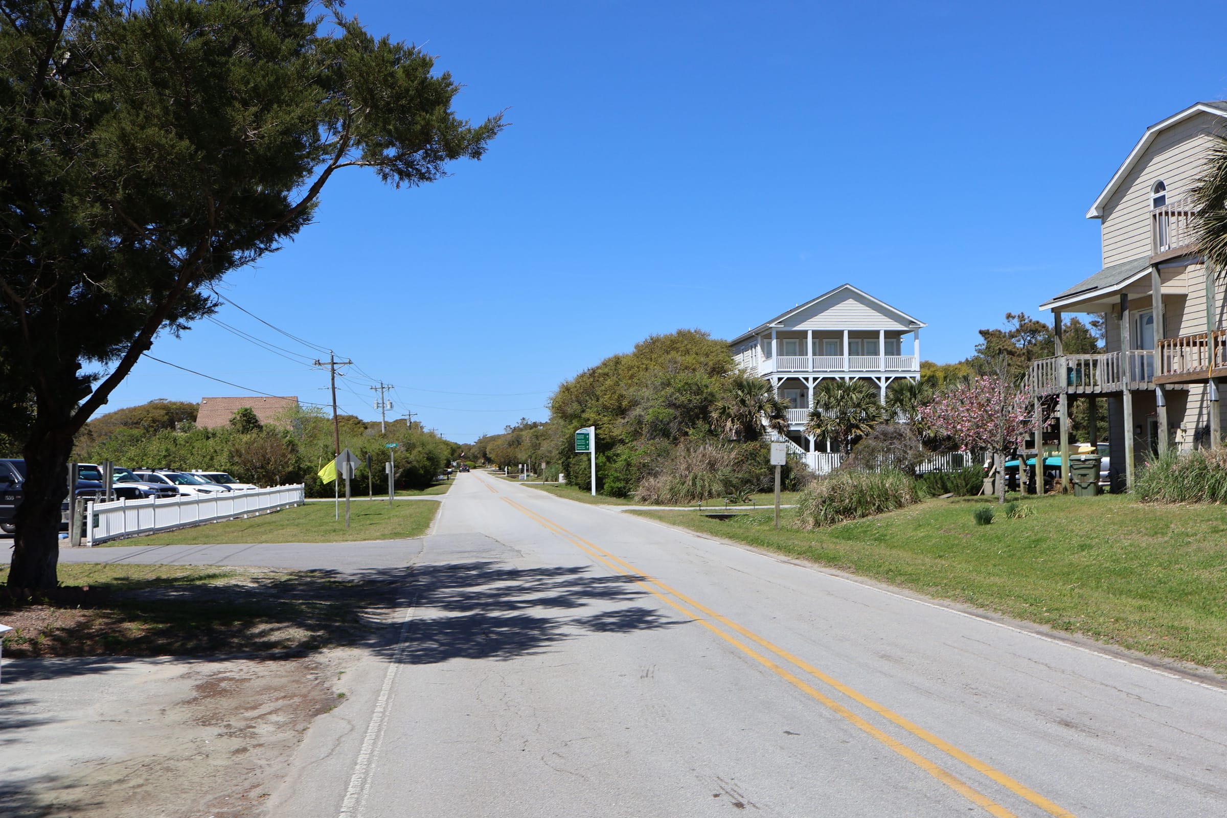 Coast Guard Road in Emerald Isle under a blue sky, raised houses with porches along the right side and live oaks on the left