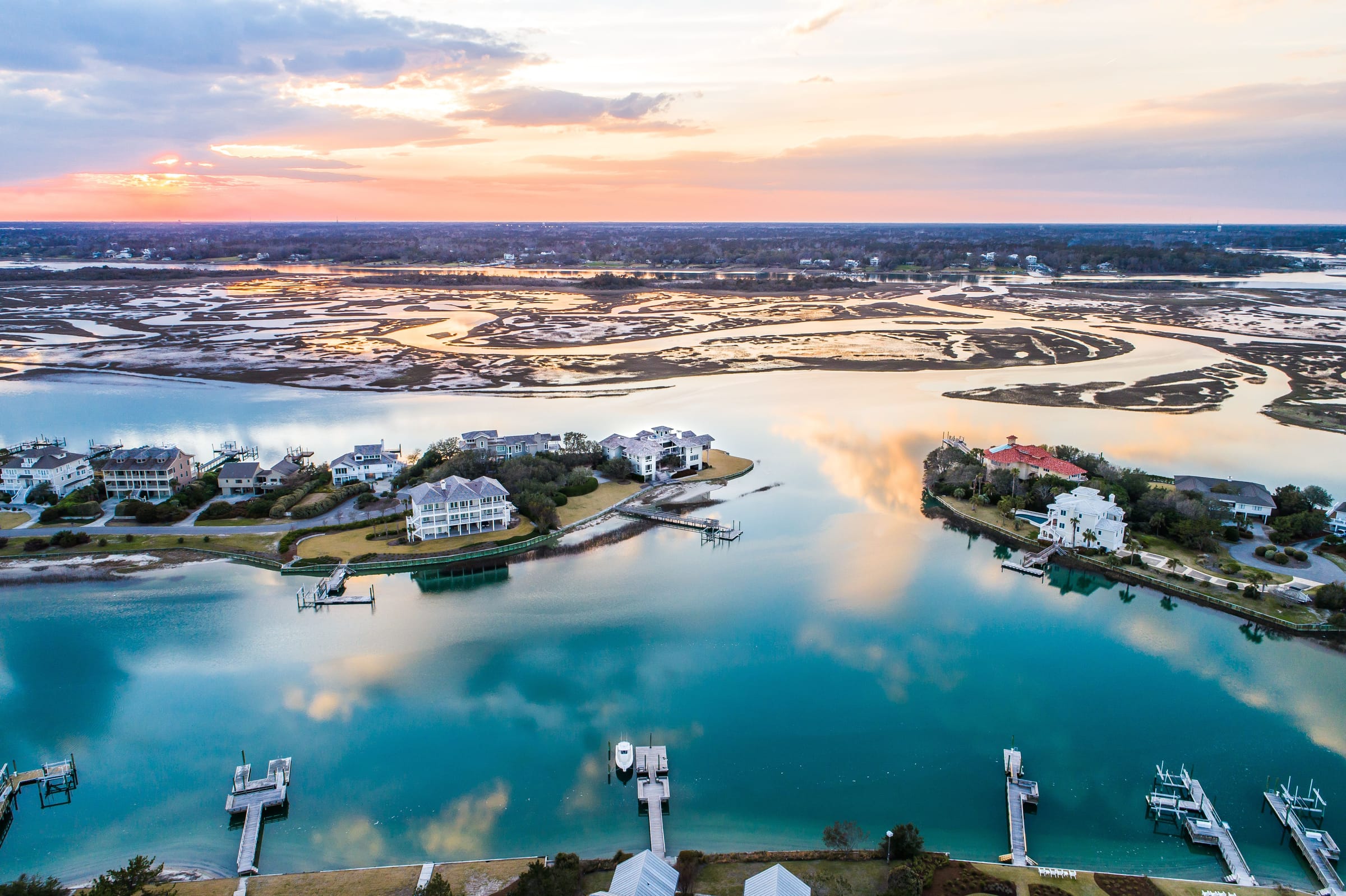 Aerial view at sunset over the waterway behind Figure Eight Island: large homes with docks on small points of land, marsh and tidal flats reflecting an orange sky