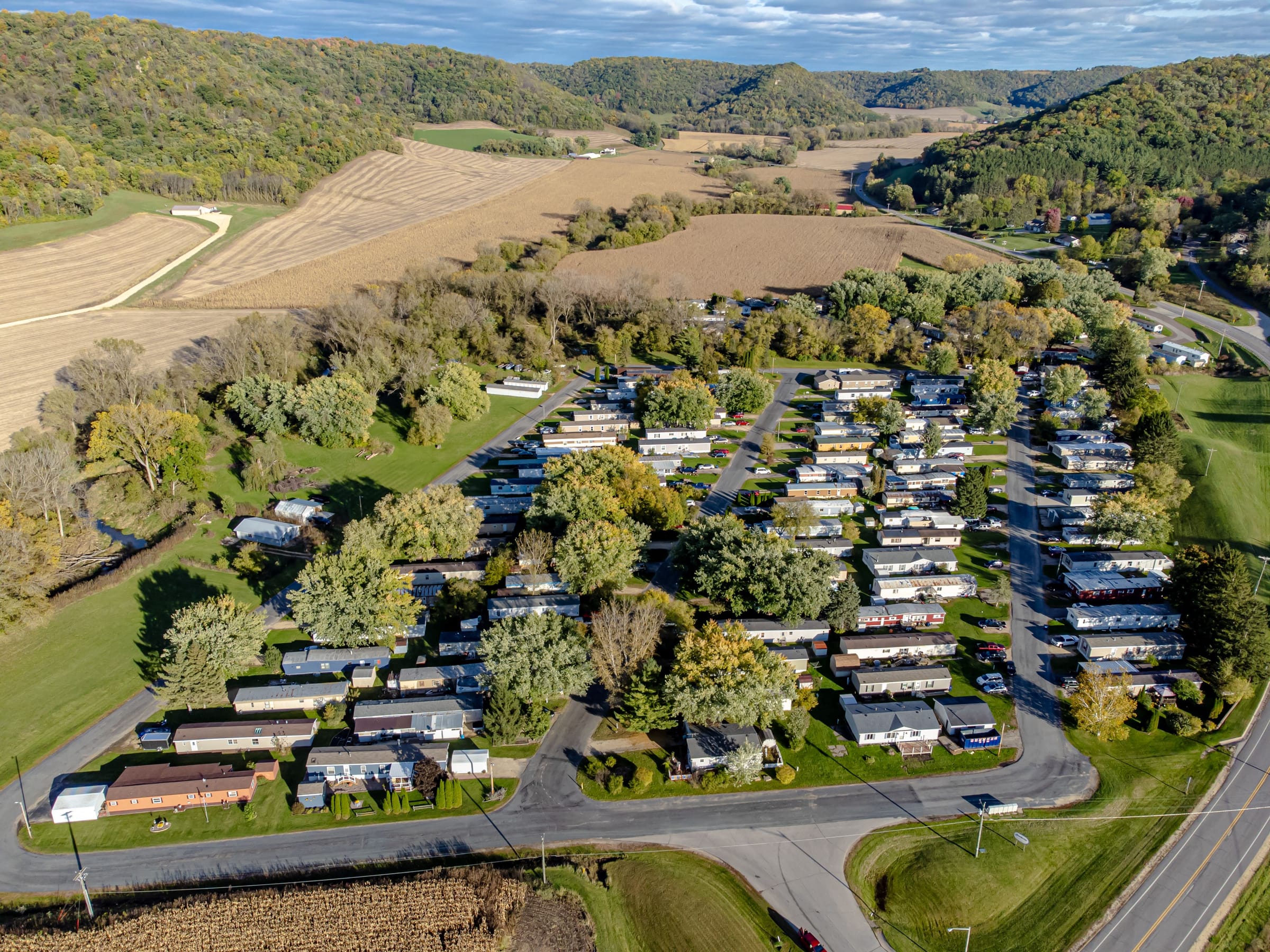 A manufactured-home community seen from the air: rows of homes along curving streets among trees, farm fields and wooded hills beyond