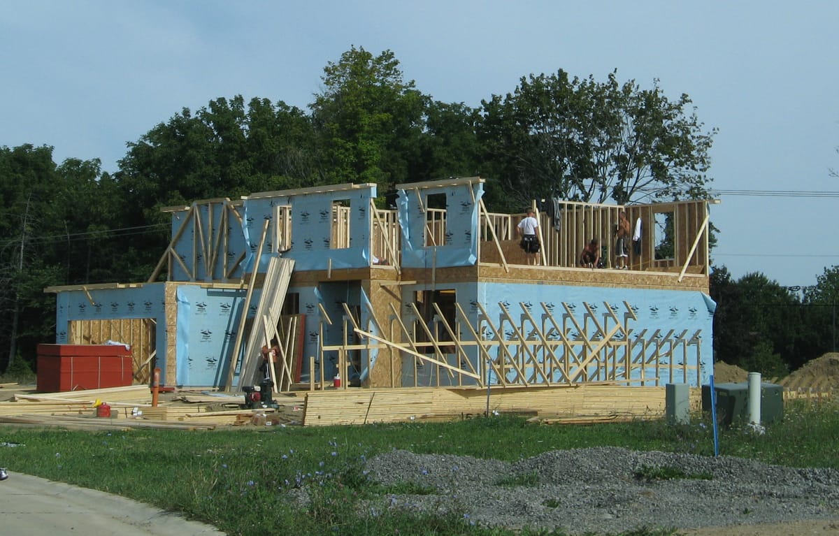 A new house being framed in a subdivision: lumber walls, blue sheathing and roof trusses going up on a cleared lot