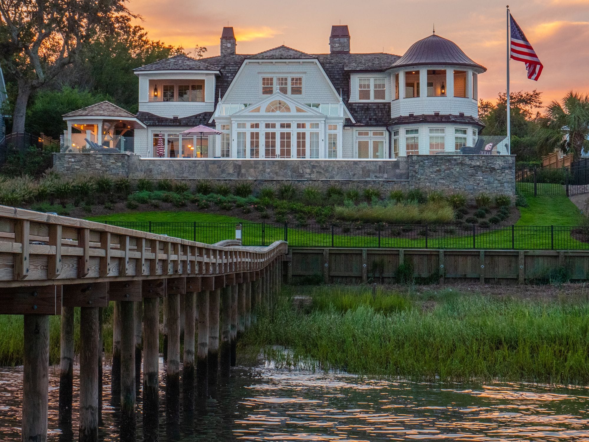 A large shingle-style waterfront home with a turret and tall windows, a flagpole, a stone wall and a wooden dock over the marsh at Wilmington