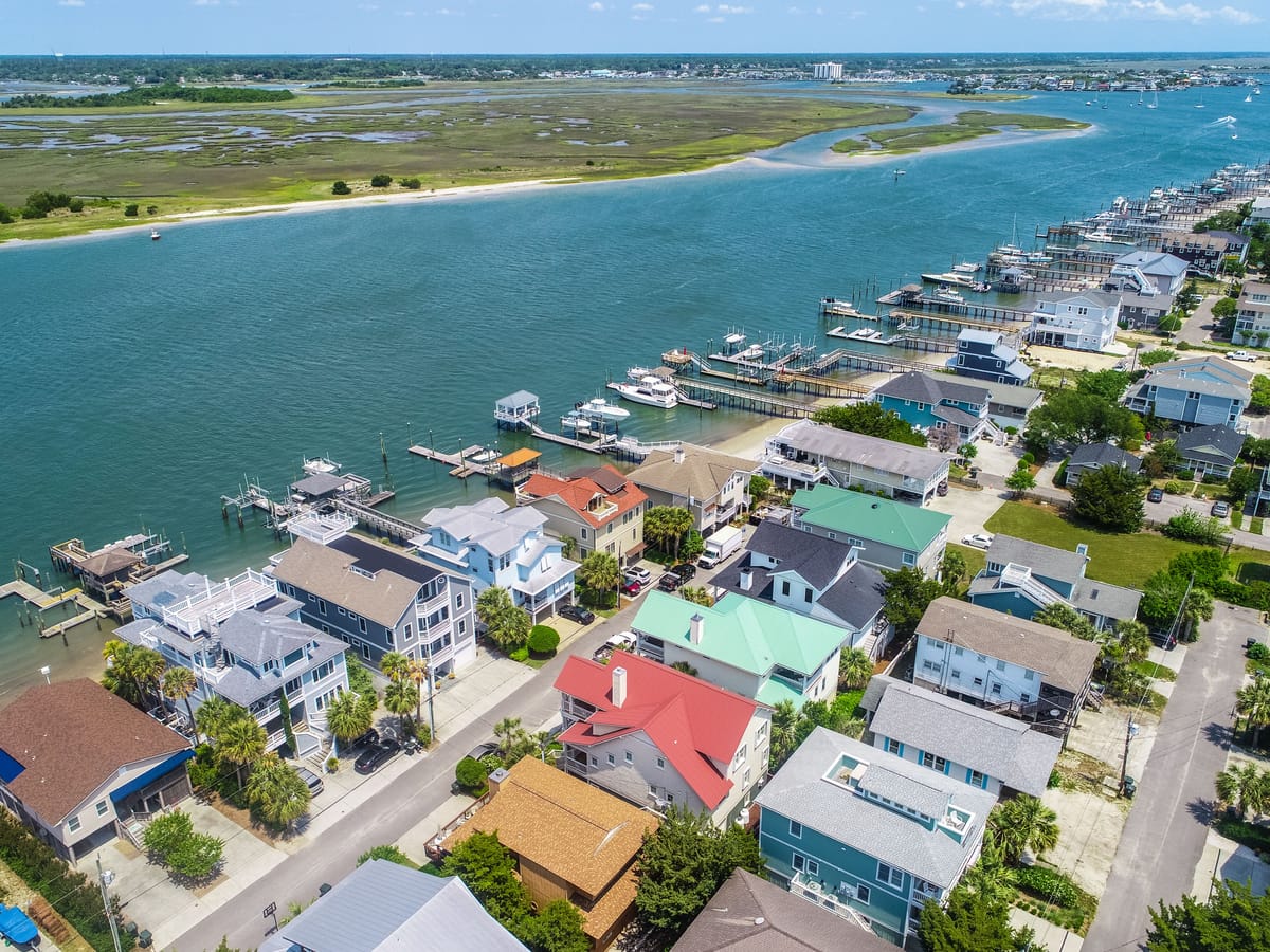 Aerial view of a Wrightsville Beach neighborhood: raised houses in white, blue and red with docks and boats on the sound side, the Intracoastal Waterway beyond